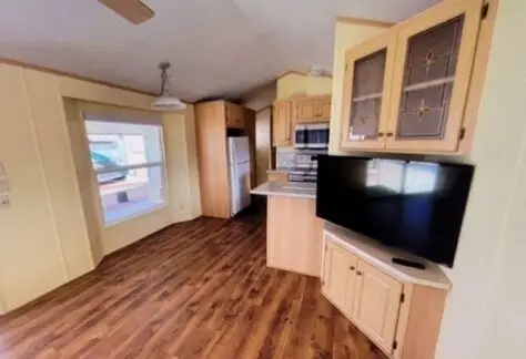A kitchen and living area with wooden floors, light wood cabinets, and modern appliances including a refrigerator, stove, and microwave. A flat-screen TV is on a corner stand. Lot 25 offers a large window on the left that brightens the space in this charming Cavco home from 2000.