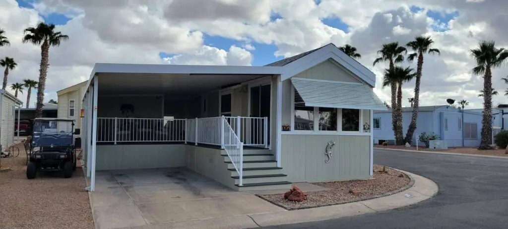 A mobile home, identified as Lot 25, with a covered porch and steps, is gracefully situated on a corner lot. Encircled by palm trees against a cloudy sky, the scene captures peace. To the left rests a golf cart, complementing this serene Cavco setting.