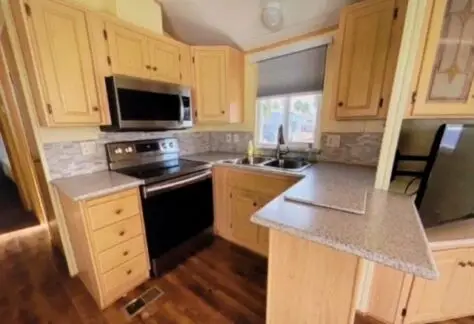 This compact kitchen in Lot 25 boasts Cavco's wooden cabinets, stainless steel appliances, and a small L-shaped countertop. Light brown flooring and a window over the sink create a naturally bright space.