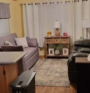 A cozy living room in Lot 25 features a gray sofa, two armchairs, and a small wooden table on the rug. A ceiling fan with lights gently illuminates the space. The light yellow walls complement the window curtains perfectly, adding charm to this 2000 Cavco home.