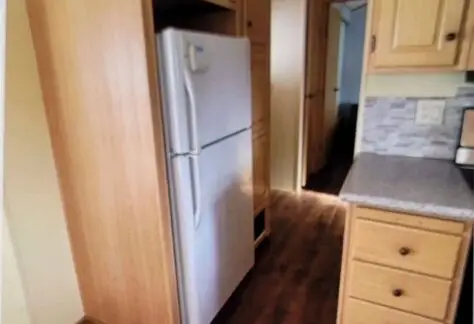 A charming kitchen with Cavco wooden cabinets, a white refrigerator, and a tiled backsplash. The wooden floor complements the rustic elegance, while the doorway marked Lot 25 beckons to another inviting room.