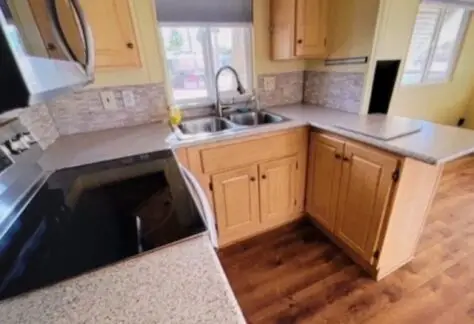Lot 25 features a charming kitchen with Cavco wooden cabinets, a stainless steel electric stove, a double sink, and light-colored countertops. Natural light streams through the window with blinds, creating an inviting atmosphere.
