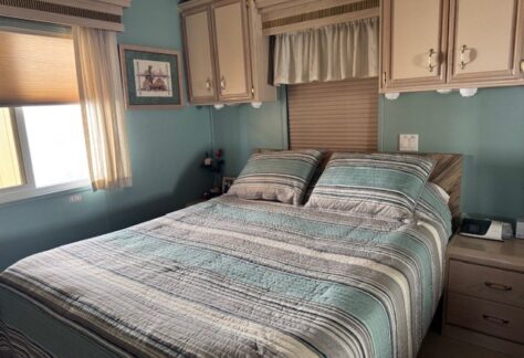 A neatly made bed with striped bedding in a cozy room with teal walls exudes charm. Above the bed, framed art and cabinets add character. A window with a shade allows natural light to dance in this inviting space by Cavco, reminiscent of Lot 316’s style from 1993.
