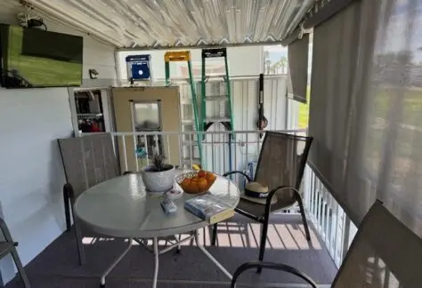 A covered balcony featuring a round table adorned with books, snacks, and a fruit bowl offers a cozy nook in Lot 316. With chairs to lounge on and a mounted TV on the wall, this charming space epitomizes the inviting warmth of the classic 1993 Cavco design.