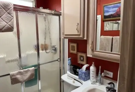 This 1993-themed bathroom, reminiscent of Lot 316 in a classic Cavco home, features striking red walls, a glass shower door, and a white sink adorned with toiletries. Cabinets provide storage beneath a stylish mirror. Towels hang neatly on racks beside the window and framed pictures.