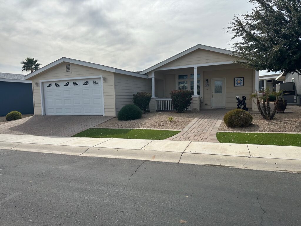 Single-story beige house with a two-car garage, white trim, and a front porch. Desert landscaping features gravel, shrubs, and pavers. Cloudy sky above.