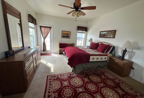 Bedroom with a wooden bed, red bedding, a dresser, nightstand, ceiling fan, and a patterned rug on a beige carpet. Two windows and a door with sheer curtains allow natural light.
