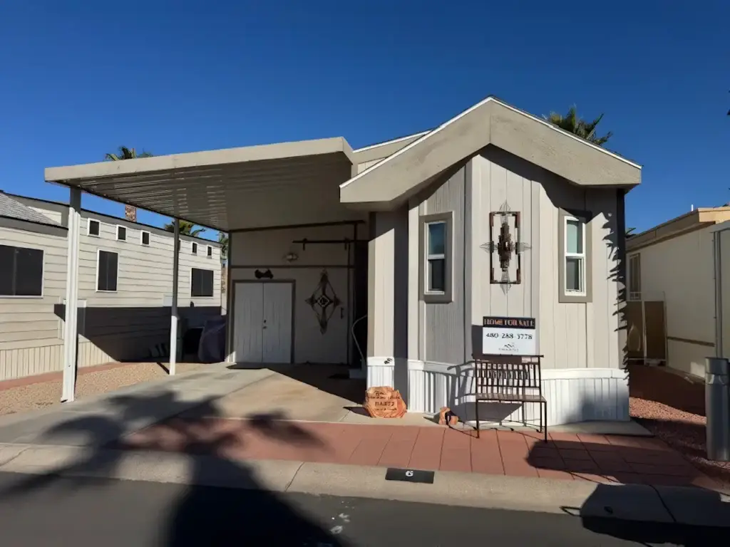 The charming Chariot Eagle mobile home, with beige siding and a covered carport, features decorative touches near the entrance like a bench and a wall hanging. A "For Sale" sign invites you to make it yours.