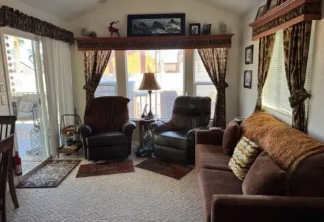 Cozy living room in a 2009 Chariot Eagle mobile home, featuring two armchairs, a couch, and a lamp near large windows. Decor includes curtains, photos, and wall-mounted items. Sunlight filters through the glass doors, enhancing the warm ambiance.