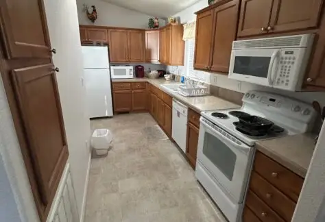 A narrow kitchen with wooden cabinets, white appliances including a stove, microwave, and refrigerator. Countertops have various items, and a trash can sits on the floor.