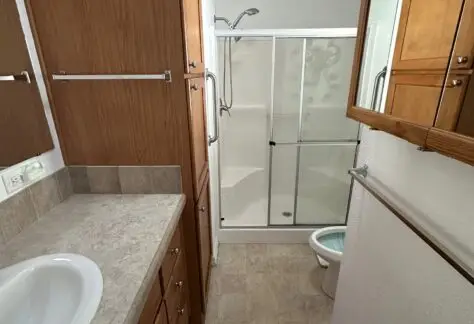 Narrow bathroom with wooden cabinets, a sink, mirror, and a glass-enclosed shower next to a toilet.