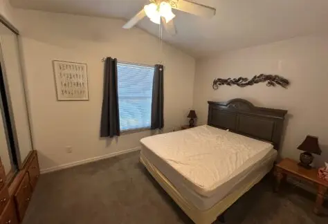 A bedroom with a bed, two side tables with lamps, and a decorative wall piece. There is a window with curtains and a ceiling fan above.
