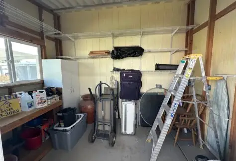 A storage room with a ladder, dolly, shelves, cabinet, boxes, and various tools and supplies. A suitcase and rolled items are also present.