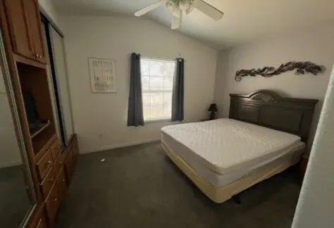 A bedroom with a double bed, a ceiling fan, and a window with gray curtains. A wooden dresser with a TV is on the left, and a decorative piece hangs above the bed's headboard.