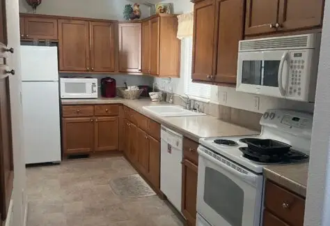 A narrow kitchen with brown wooden cabinets, light countertops, a white stove, microwave, and refrigerator. Rooster decorations are above the cabinets.