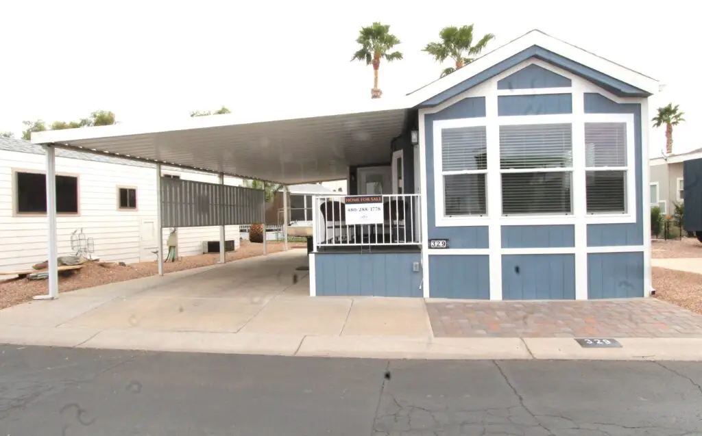 A blue and white Cavco manufactured home sits on Lot 329, featuring a covered carport, stone driveway, and inviting porch. In the background, two palm trees sway gently in the breeze.