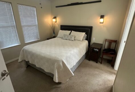 Bedroom with beige walls, a bed with white bedding, two wall-mounted lamps, and a ceiling fan. Two windows with blinds are on the left, and there is a small nightstand with a chair nearby.