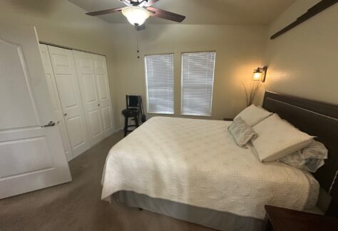 Bedroom with a large bed, white bedding, two windows with blinds, a ceiling fan, closed closet doors, and a small chair by the corner.