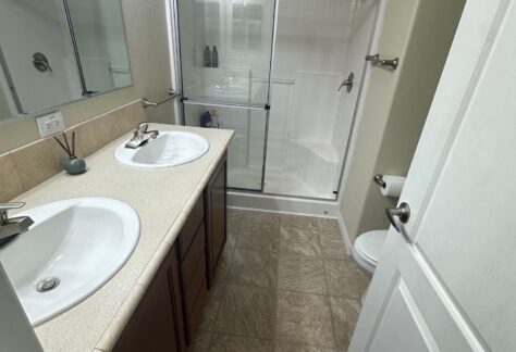 Bathroom with dual sinks, mirror, and a glass-enclosed shower. Light fixtures are above the mirror, and there's a closed door on the right. The floor is tiled.