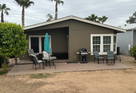 Single-story house with patio furniture, a grill, and an umbrella on a paver patio. Palm trees and shrubs surround the area under a cloudy sky.