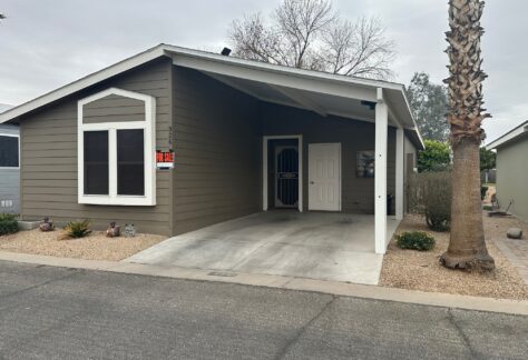 Single-story house with a carport and 'For Sale' sign. Features include a concrete driveway, light brown siding, white trim, and desert landscaping with small plants and a palm tree.