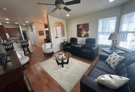 A cozy living room with a ceiling fan, two recliners, a sofa, a coffee table, and a rug on wooden floors. Kitchen is visible in the background. Bright window lighting.