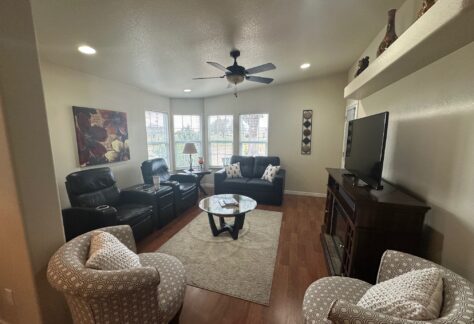 Living room with two leather recliners, two patterned armchairs, a wooden TV stand, and a ceiling fan. The room has a round coffee table and is decorated with paintings and small sculptures.