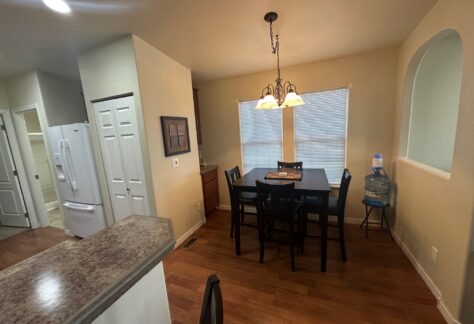 Dining area with wooden table and four chairs, lit by a hanging light fixture. Nearby, a water dispenser stands by the windows with blinds. Adjacent kitchen with visible refrigerator.