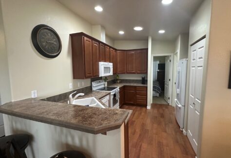 Kitchen with wooden cabinets, marble countertops, and white appliances. Includes a stove, microwave, and refrigerator. Hardwood floor and a decorative clock on the wall.