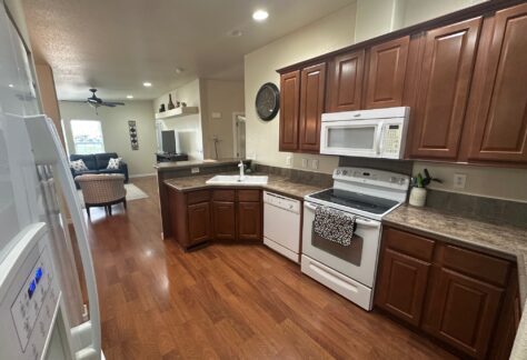 Kitchen with wooden cabinets, white appliances, and brown countertops. Open layout leads to a living room with a ceiling fan, TV, and seating area in the background.