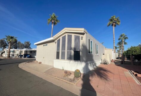 A mobile home with a small garden area in front, situated on a paved road, with palm trees and a clear blue sky in the background.