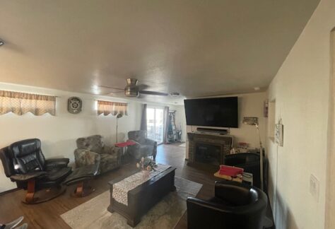 Living room with various chairs, a coffee table, ceiling fan, and a large wall-mounted TV. There's a rug on the wooden floor and a small fireplace beneath the TV.