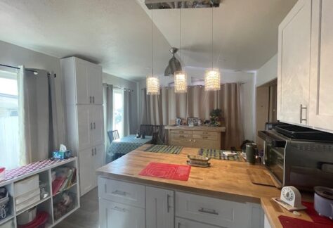 A kitchen with a wooden countertop, hanging pendant lights, appliances, storage shelves, a dining table with a green tablecloth, and a sideboard with framed photos and plants.