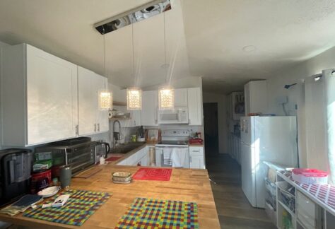 A modern kitchen with white cabinets, hanging pendant lights over a wooden island, and various appliances. Bright sunlight enters through a door on the right.
