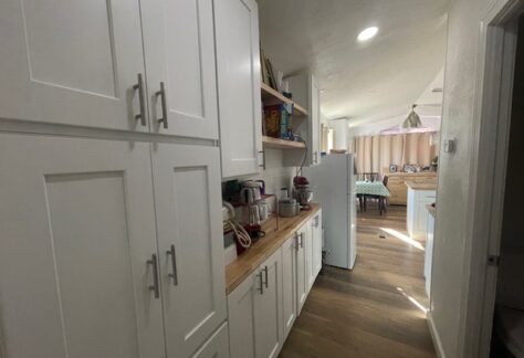 A narrow kitchen with white cabinets, wood countertops, and a refrigerator. Shelves hold small appliances and containers. Sunlight filters through the window, illuminating the space.