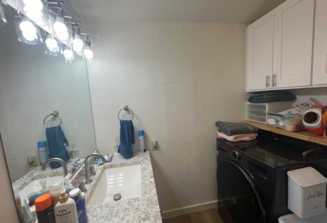 Bathroom and laundry room with a countertop sink, toiletries, and a mirror on the left. On the right, a washing machine with folded towels and storage items above.