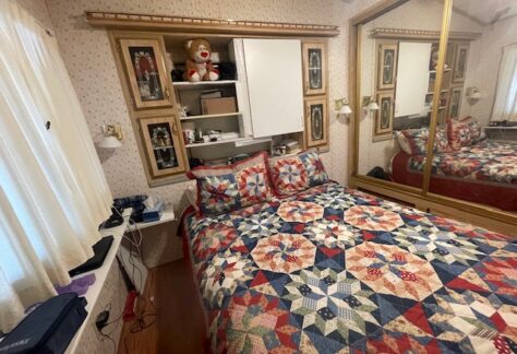 A small bedroom with a colorful quilted bed, mirrored closet, shelves with dolls and books, and a window with sheer curtains.