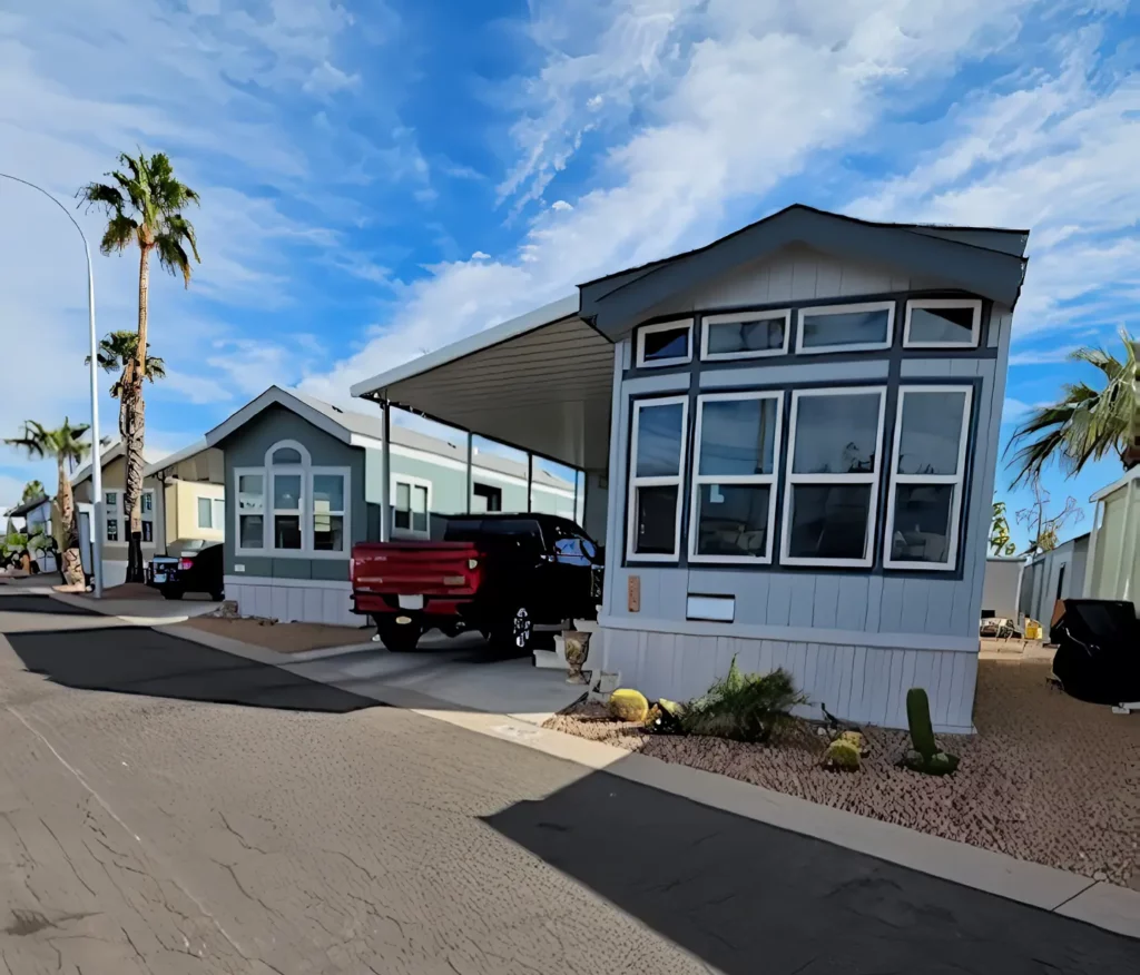 Under a partly cloudy sky, Cavco mobile homes line up neatly, each with carports. A red truck rests beside one of the 2016 models on Lot 172. Desert landscaping adds charm to the foreground.