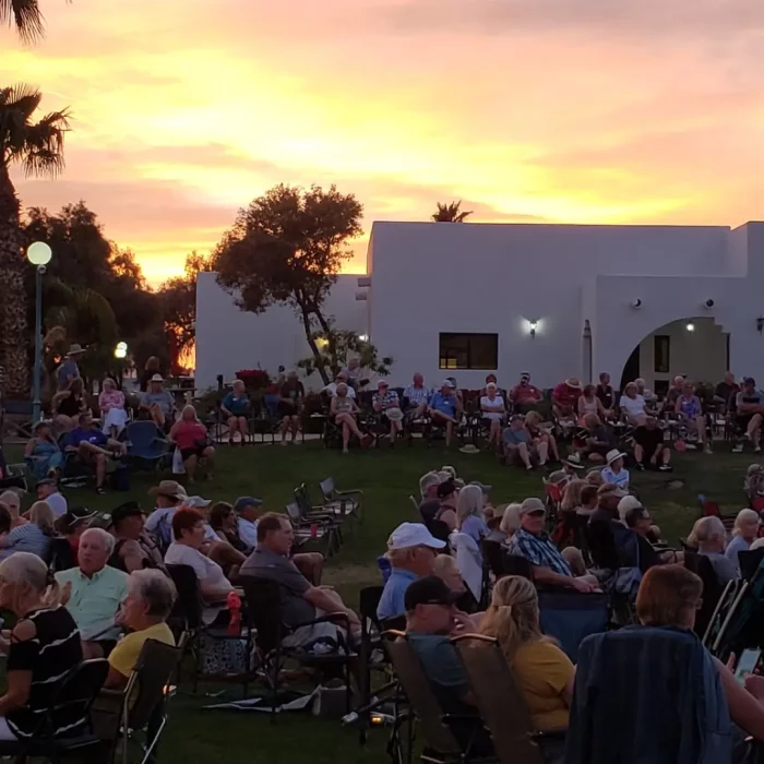 An enchanting outdoor gathering at Pueblo El Mirage unfolds as the sun sets, with people comfortably seated on chairs in a grassy area. Surrounded by trees and charming white buildings, this Arizona winter evening captures the essence of Winter Bliss.