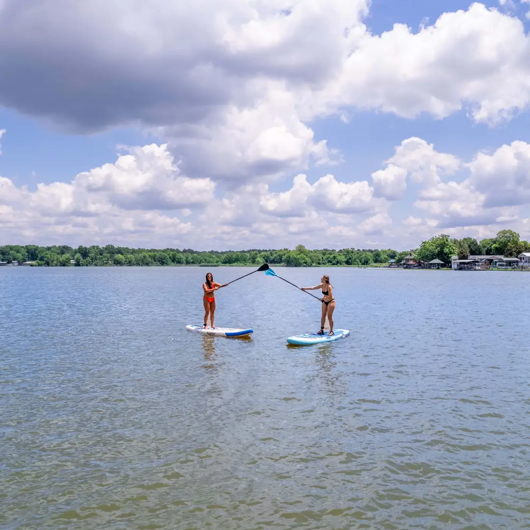 Two people on paddleboards touch their paddles together on the tranquil waters of Lake Conroe, enjoying water-based activities under a cloudy sky.