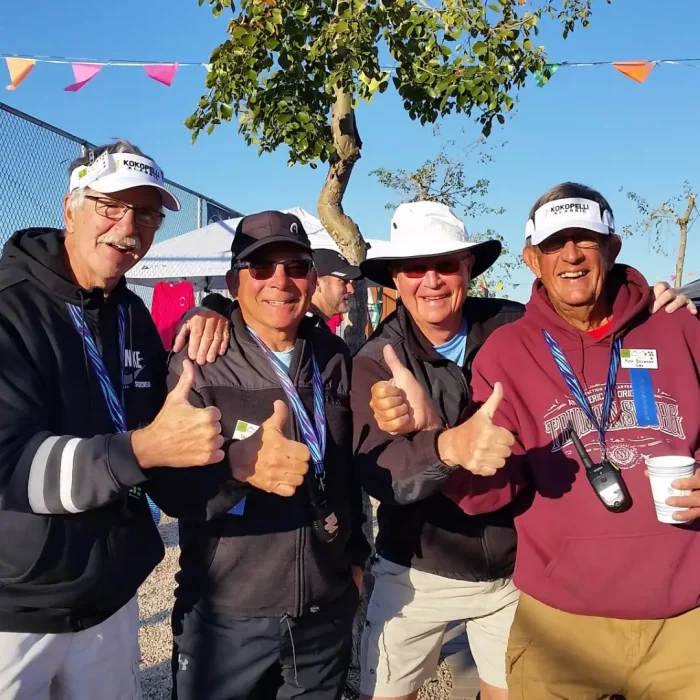 Four men, likely Arizona winter visitors, are posing outdoors, all wearing hats and sunglasses while giving a thumbs-up. They are smiling with colorful flags and a tree in the background, enjoying the benefits of an early booking for their trip.