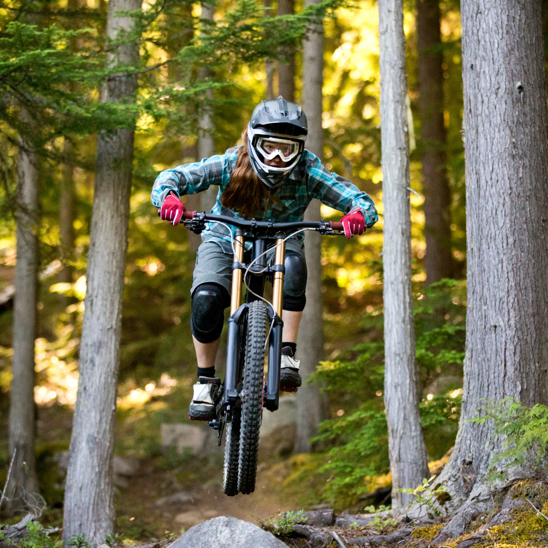 A person wearing a helmet and protective gear rides a mountain bike in Park City's forest, catching air over a rocky path.