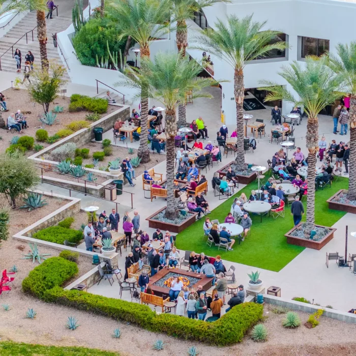 Aerial view of a busy outdoor patio in Gold Canyon, where people engage in lively activities at tables, surrounded by palm trees and lush landscaping.