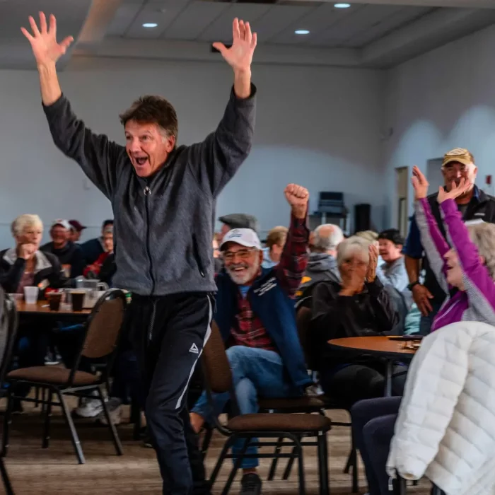 A man with raised arms celebrates energetically in a room filled with winter visitors sitting at tables, also appearing joyous.