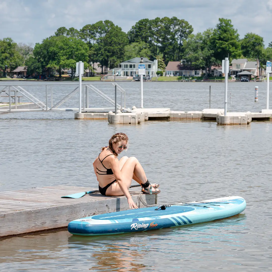 A person sits in ultimate relaxation at the water's edge on a dock beside a paddleboard, securing a strap around their ankle. The background reveals serene trees, quaint houses, and a quiet lake.