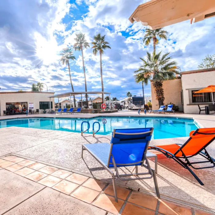 The outdoor pool area at Sunrise RV Resort offers a serene escape with lounge chairs nestled among palm trees. The beauty of the partly cloudy sky is reflected in the water, while a building stands under the bright Arizona winter sun.