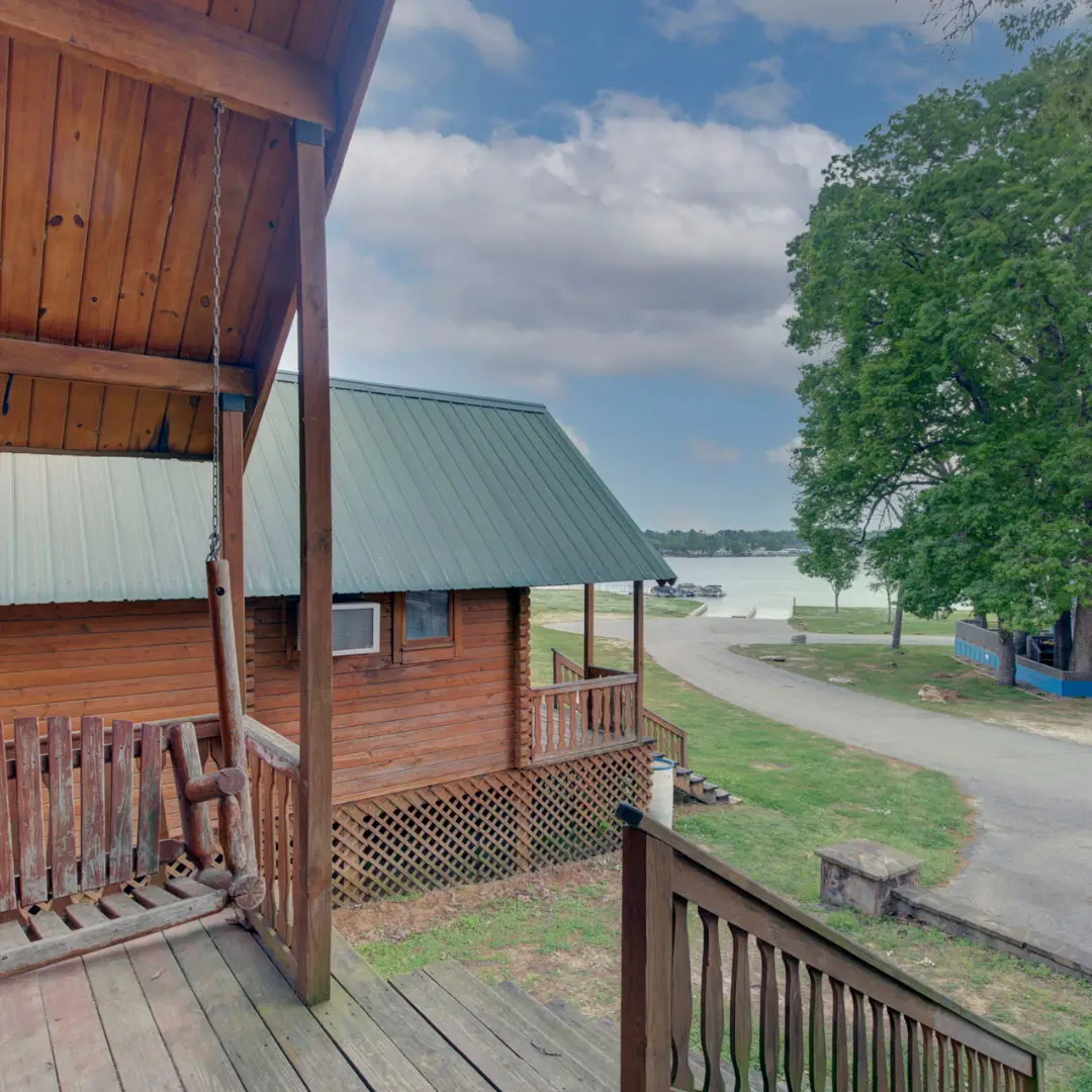 Wooden cabins with green roofs are nestled near a lake, perfect for a spring break retreat at Lake Conroe Cabins. A wooden porch swing graces the foreground, and a road curves toward the serene waters. A large tree stands on the right, inviting families to explore this idyllic setting.