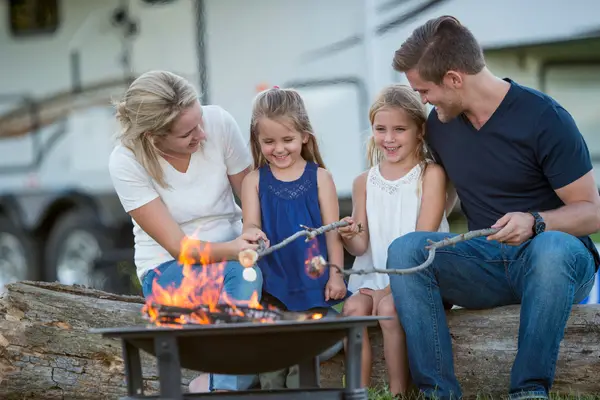 A family of four enjoys camping with kids, roasting marshmallows over a campfire while sitting on a log, with their camper cozy in the background—a perfect family camping activity.