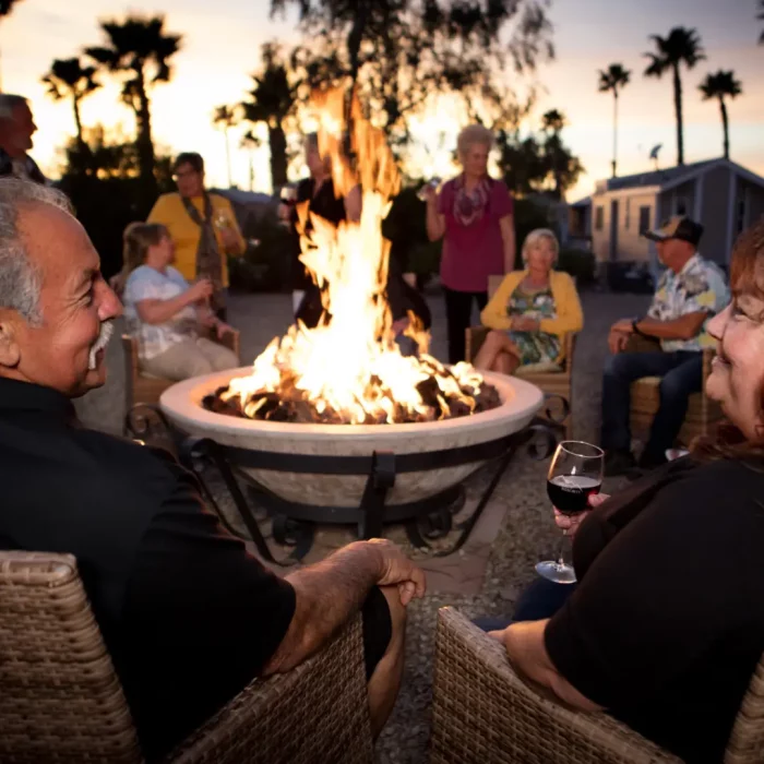 In the cool January evening, a group of people gathers around a fire pit in Gold Canyon, sitting on chairs and holding drinks. With palm trees swaying in the background, they enjoy one of their favorite activities under the starry sky.