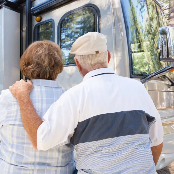 Two elderly people, seen from behind, stand close together, contemplating a large RV at a Winter RV site. The person on the left has brown hair, while the person on the right is wearing a beige cap.